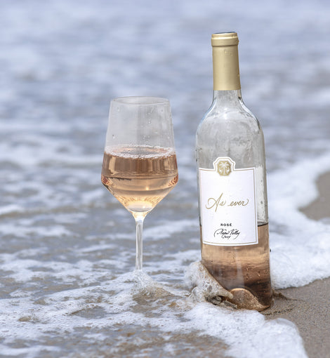 Bottle and glass of As ever Rosé wine on a beach with ocean waves in the background