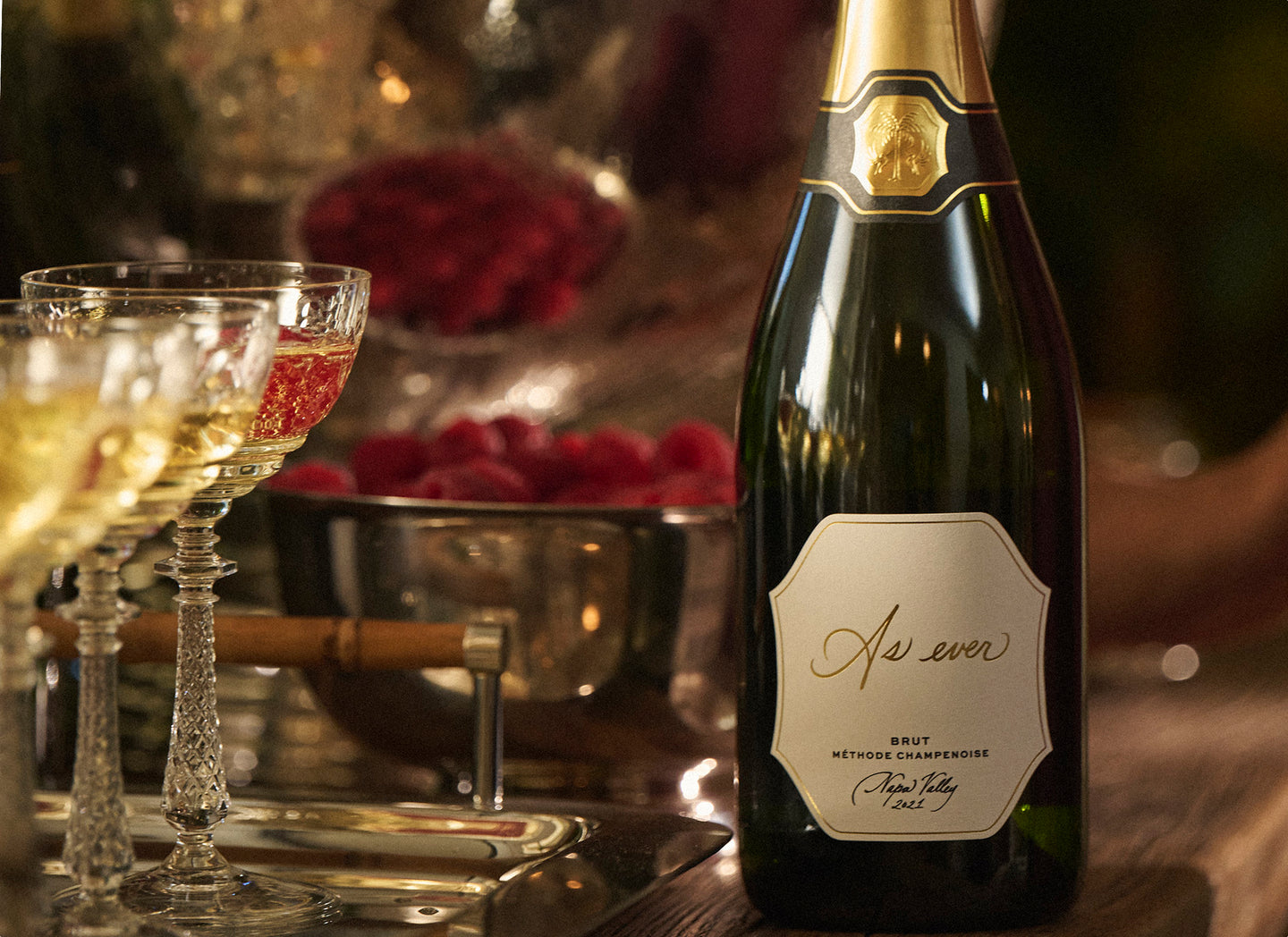 Bottle of As ever Brut with a cork on a wooden table, next to wine glasses and fruit.