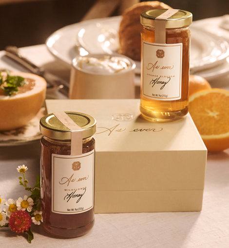 Two jars of As ever honey with a decorative label on a table with a box, flowers, and fruit.