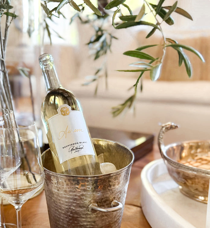 Bottle of As ever Sauvignon Blanc in a metal ice bucket with a glass on a table, surrounded by greenery