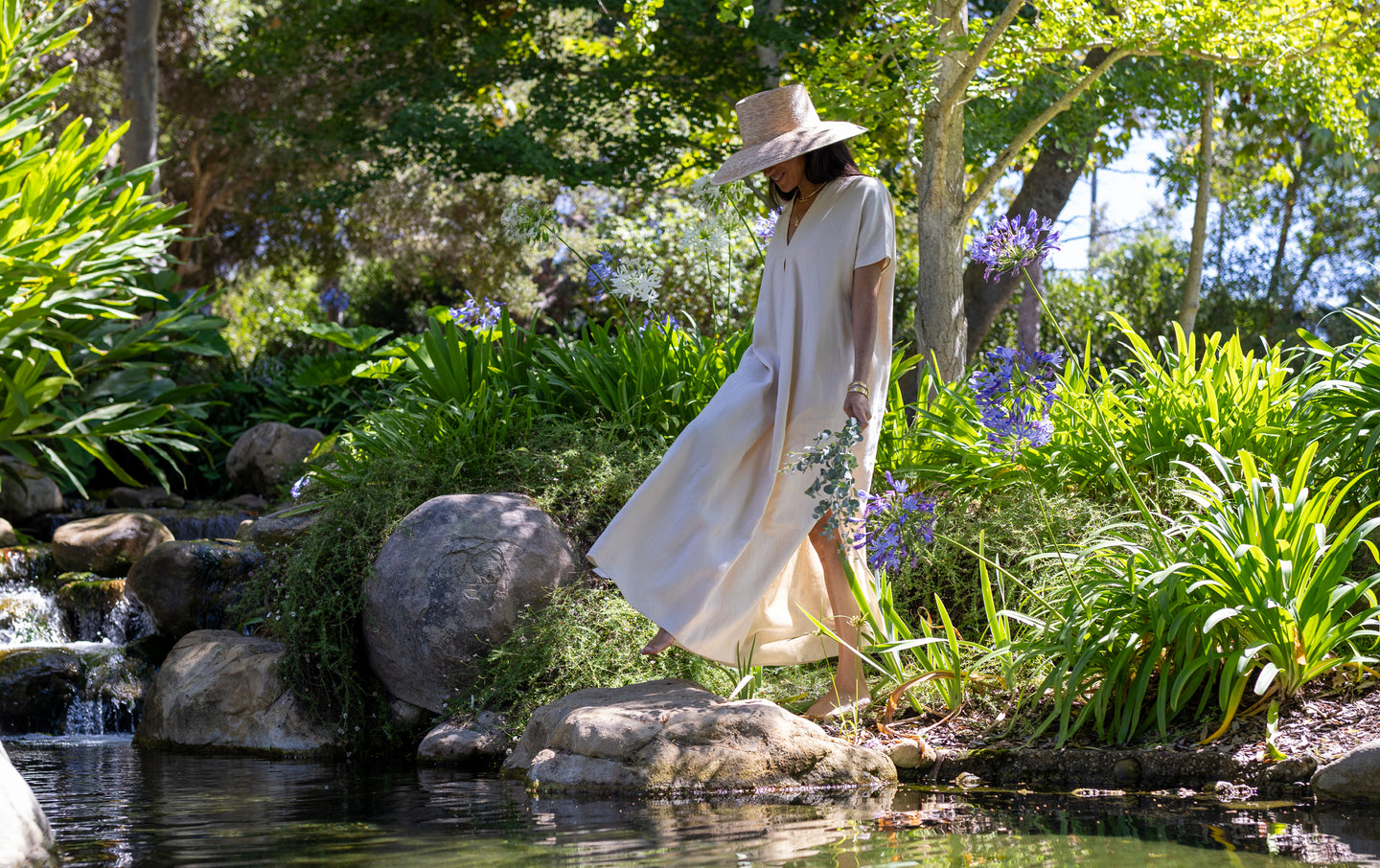 Meghan in a flowing dress and hat walking near a pond with greenery and flowers.
